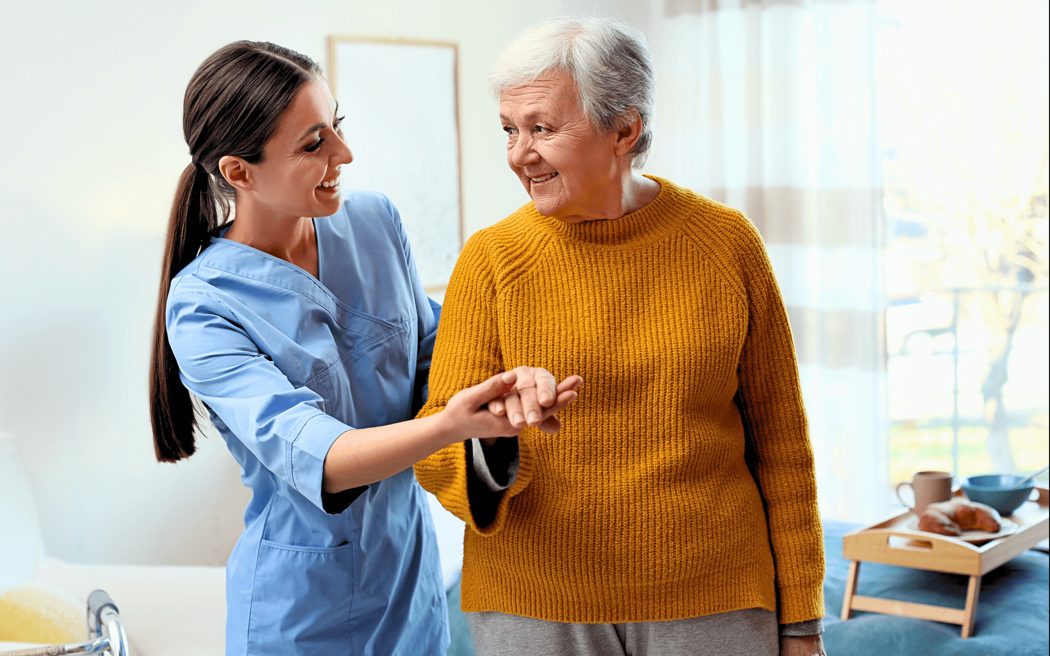 A caregiver assists an elderly woman with walking. The caregiver is smiling as the woman looks on with a smile.
