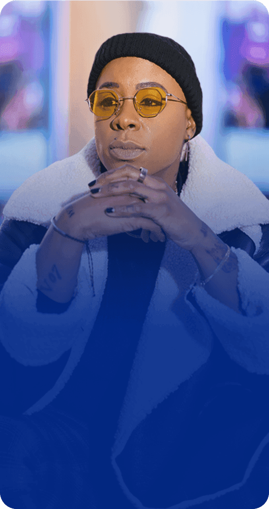 Ashleigh Warren, a person sits thoughtfully in front of multiple colorful screens, dressed in a stylish jacket.