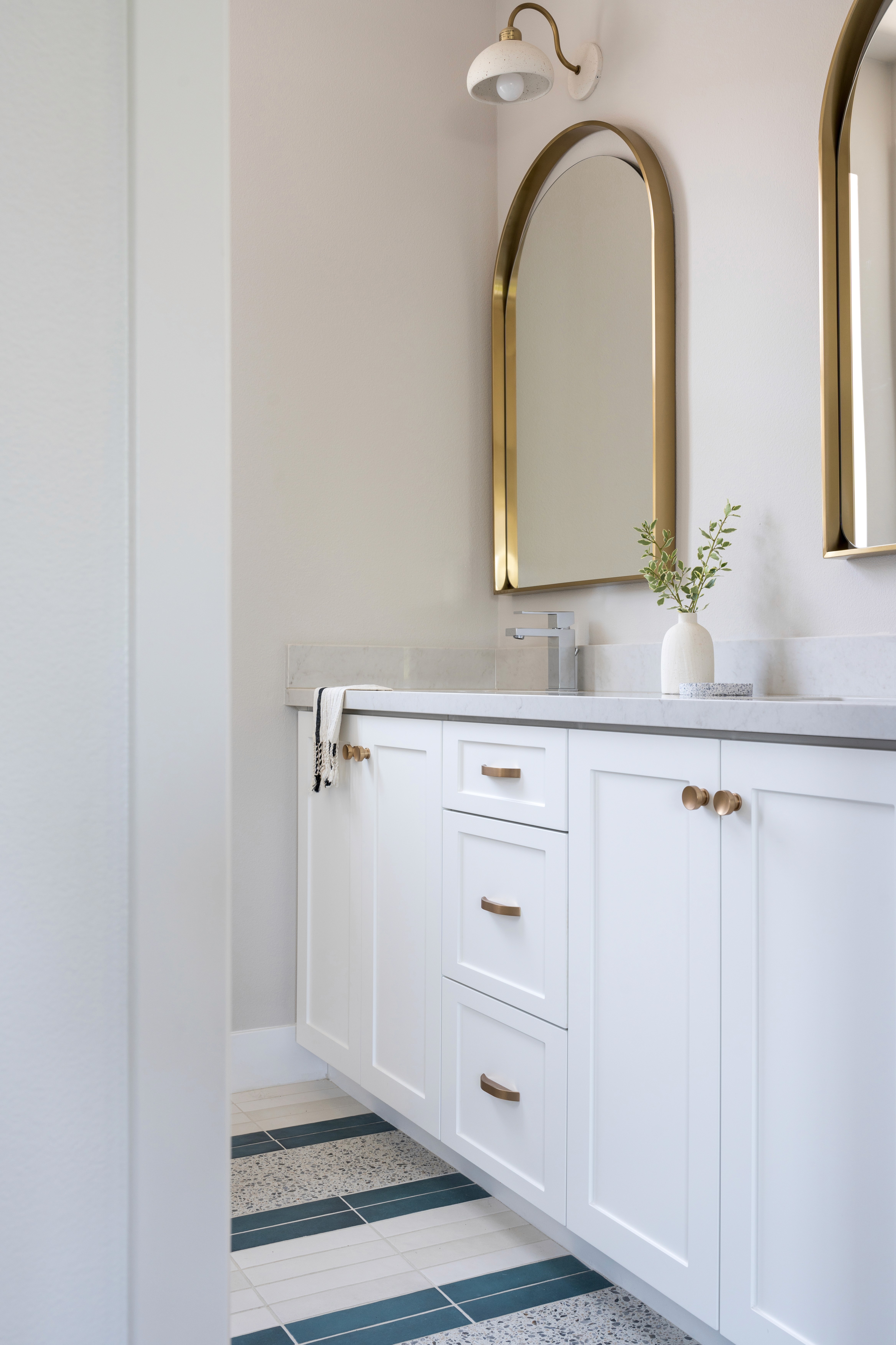 Dual vanity bathroom with arched, gold framed mirrors, white wall lamp with gold accents, grey marble countertop, white cabinetry with gold finishes, and multi-colored tile flooring by Jubilee Interiors in Arcadia, California