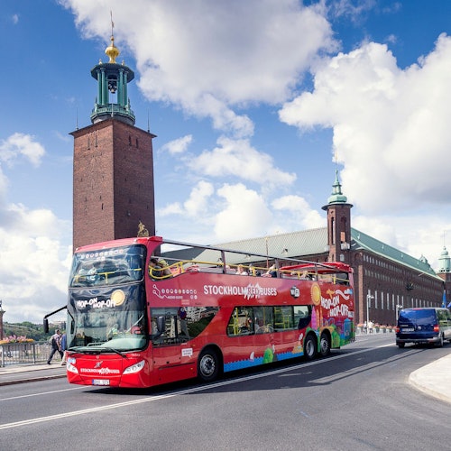 A red double-decker tour bus drives past Stockholm City Hall under a blue sky with scattered clouds.