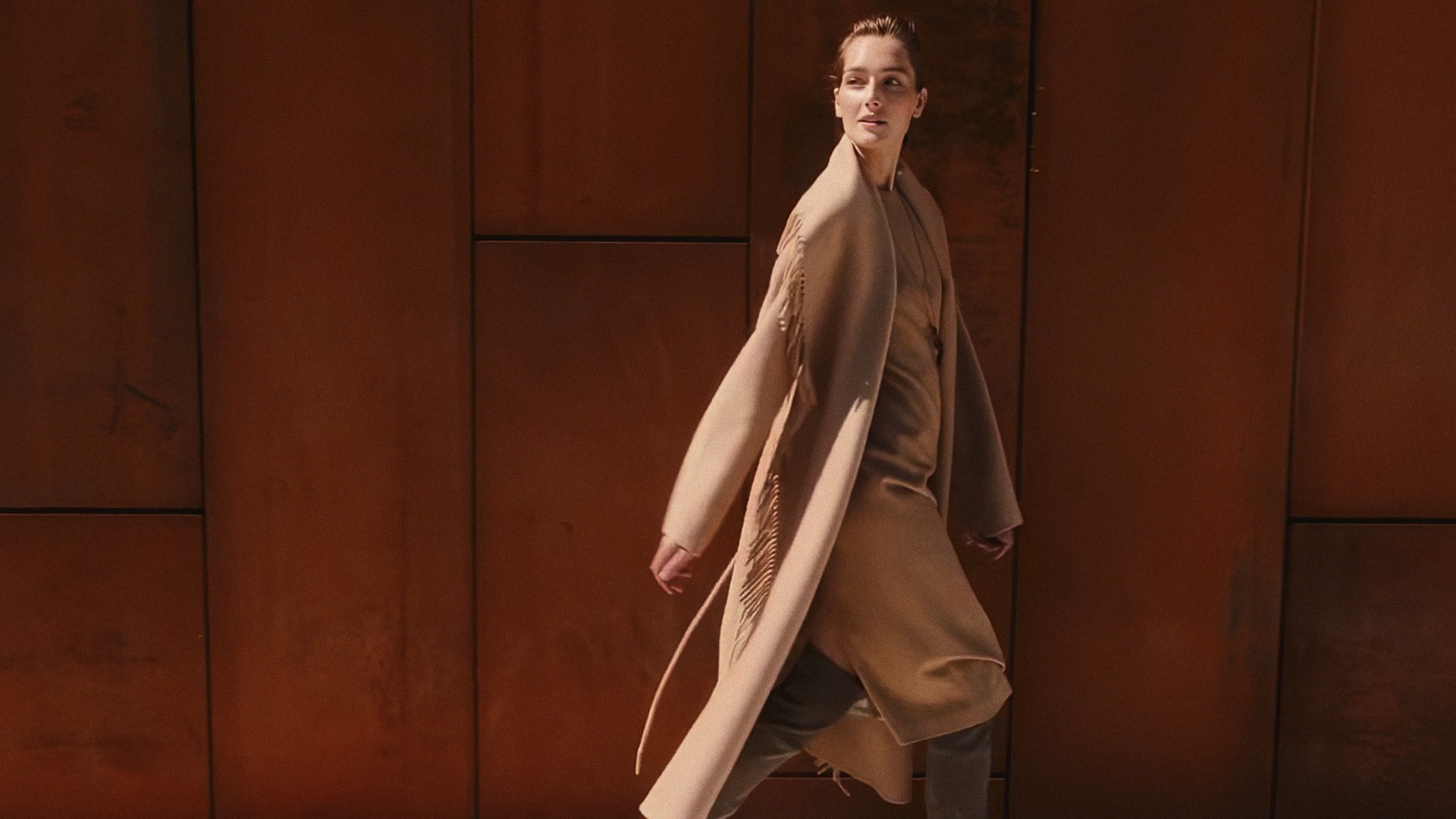 A woman walking in a long camel coat against a rust-colored steel wall, Lafayette 148 New York