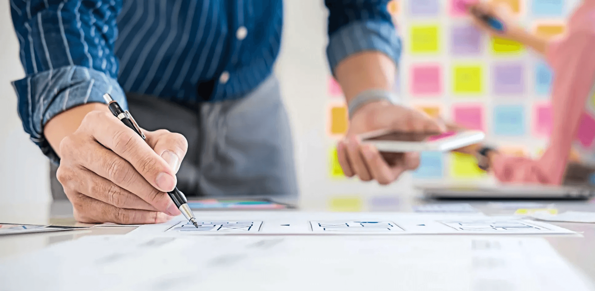 Person zeichnet mit einem Stift ein Diagramm auf Papier, während sie ein Smartphone hält; im Hintergrund bunte Haftnotizen an einer Wand für Planung oder Ideenentwicklung.