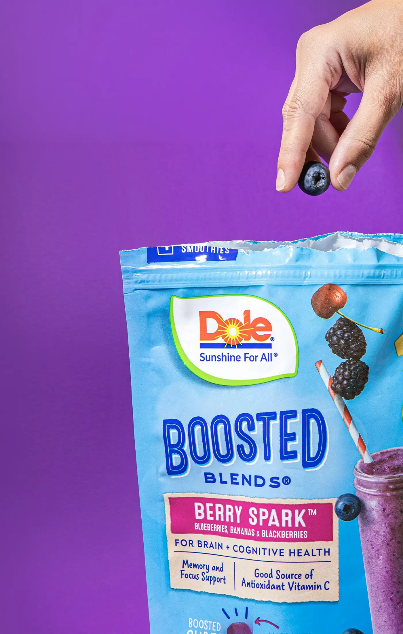 A vibrant bag of Dole Boosted Blends Berry Spark smoothie mix on a purple background. A hand holds a blueberry above the open bag, evoking a fresh and healthy vibe.