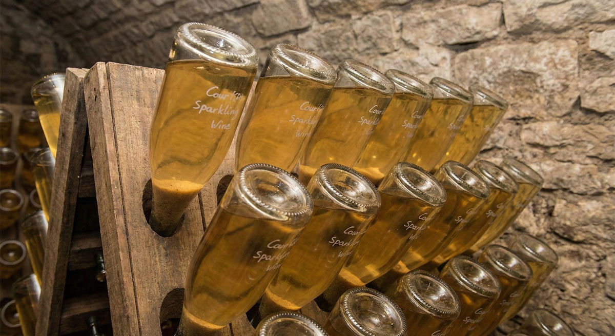 Close-up of sparkling wine bottles resting neck-down in a traditional wooden riddling rack within a stone cellar. The clear glass reveals golden wine with yeast sediment collected in the necks, illustrating the final stages of the traditional bottle fermentation process.