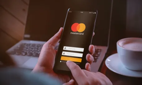 Hände halten ein Smartphone mit dem Mastercard-Anmeldebildschirm. Im unscharfen Hintergrund Laptop und Tasse auf einem Tisch. Warmes Licht.
