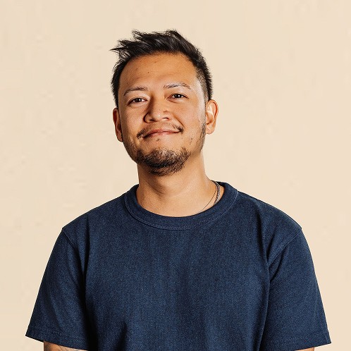 Man with short hair and light facial hair wearing a dark T-shirt, standing against a neutral background.