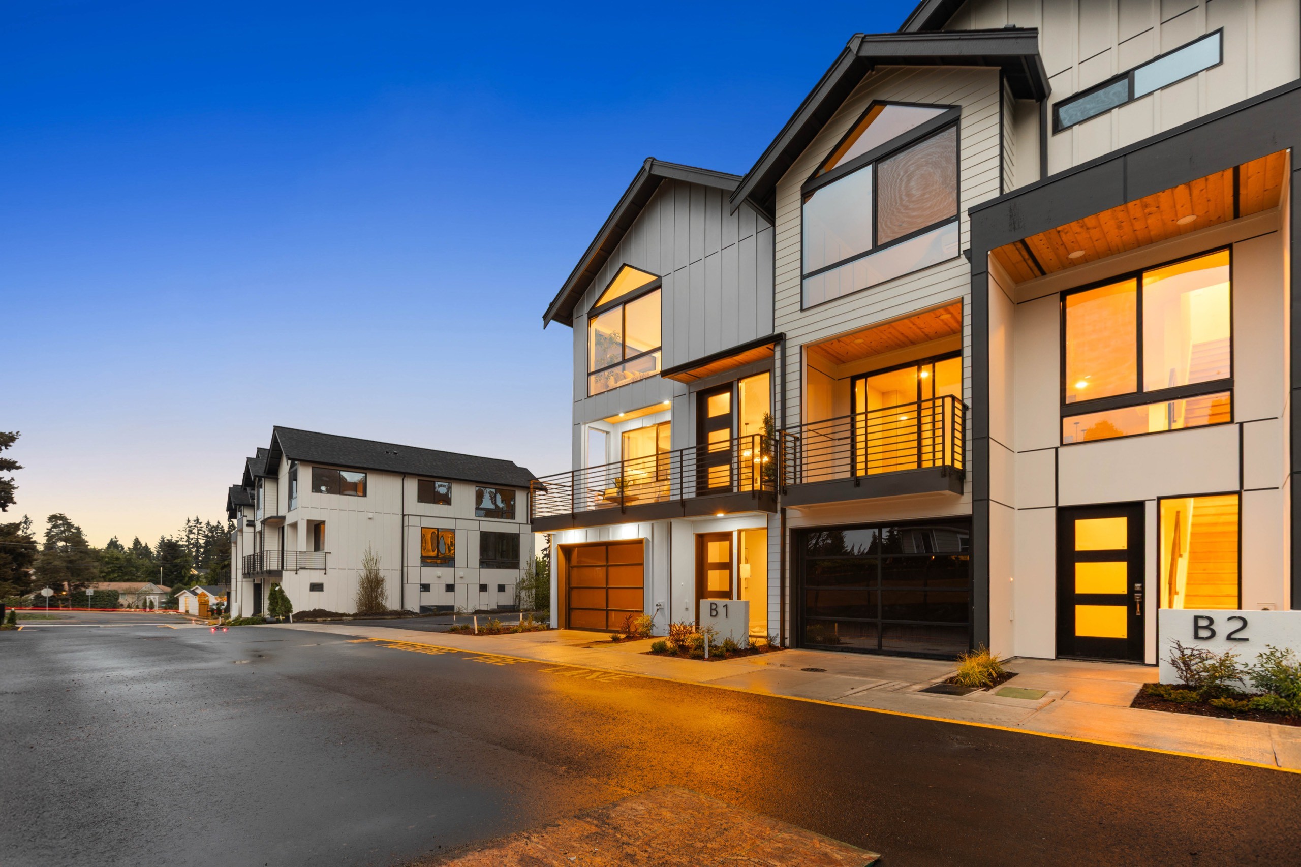Dusk street-level view of a newly built luxury townhome with two-car garage, large balcony, floor-to-ceiling windows, and contemporary grey and white exterior