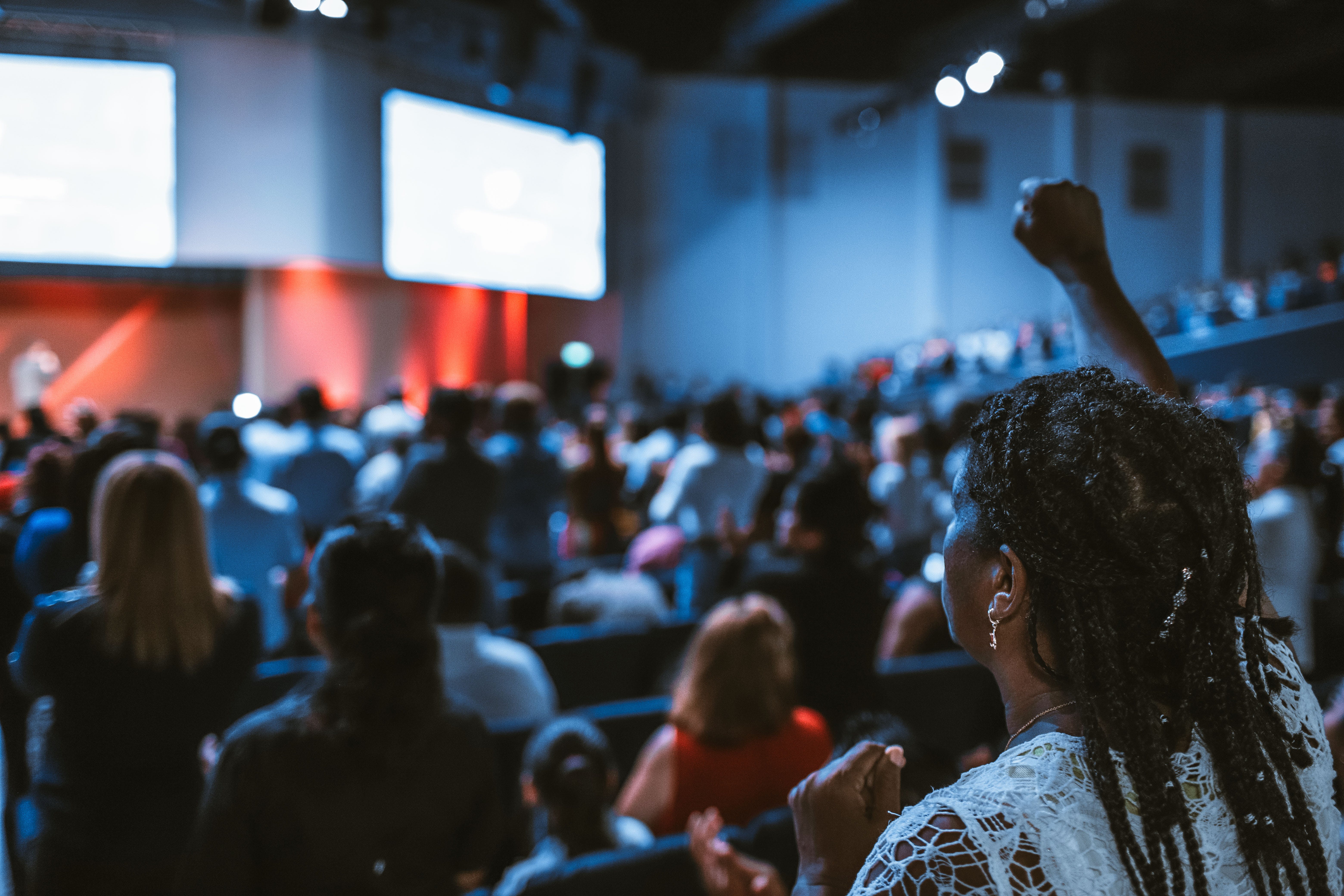 A woman raises her hand in front of a large crowd, wanting to contribute.