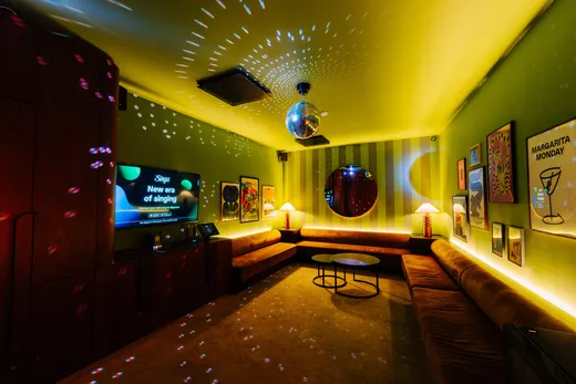 A young person with curly hair smiles at the camera while sitting in front of a laptop against a dark background.