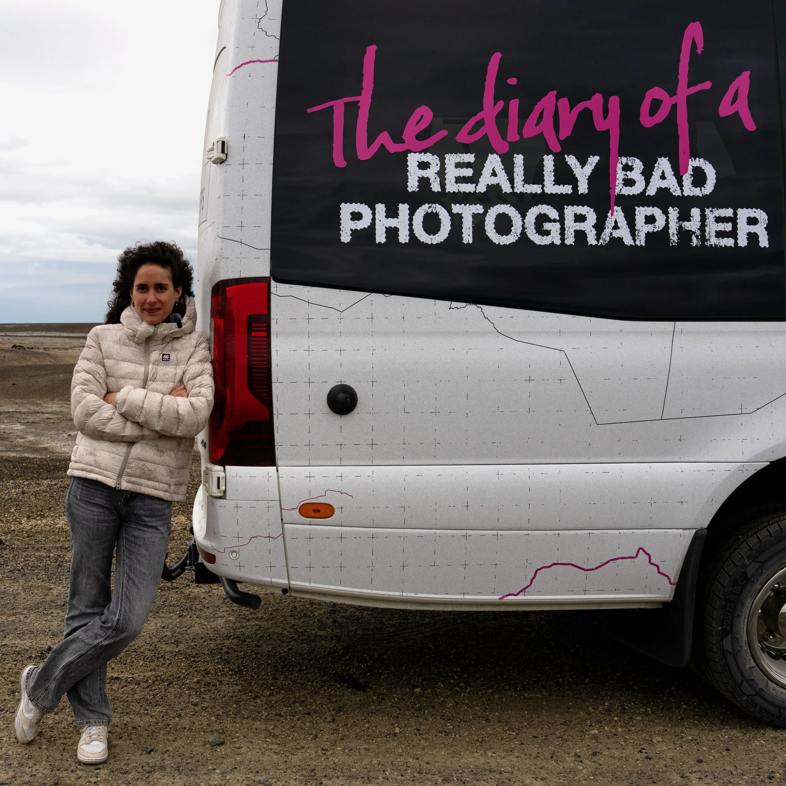 Blanche Mortemard stands in front of the Icelandair "Diary of a Really Bad Photographer" tour bus in the Icelandic countryside, its side panel reading "Caution: A really bad photographer may emerge, pose at own risk.