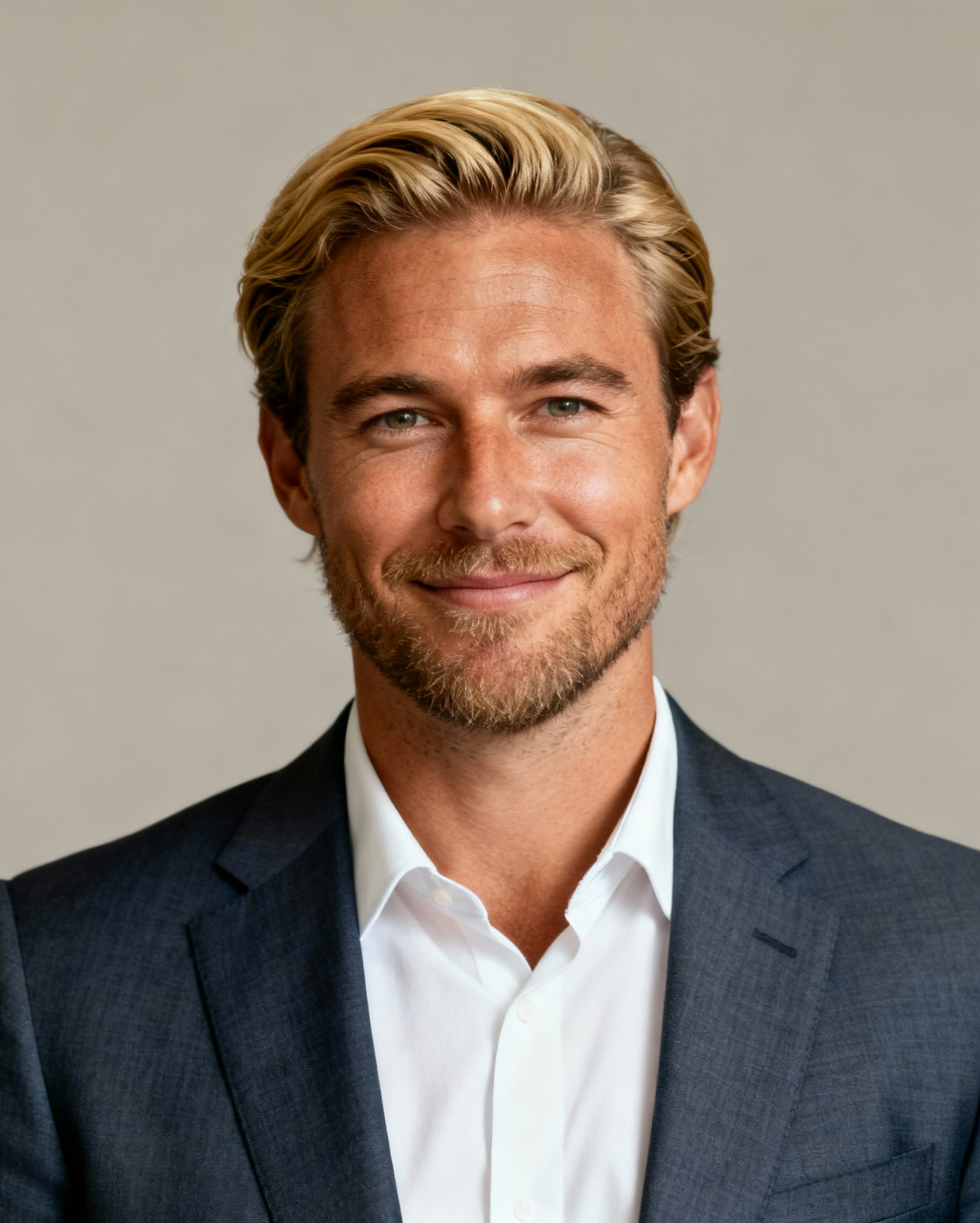 A smiling man with blonde hair and a beard, dressed in a suit and white shirt, against a neutral background.