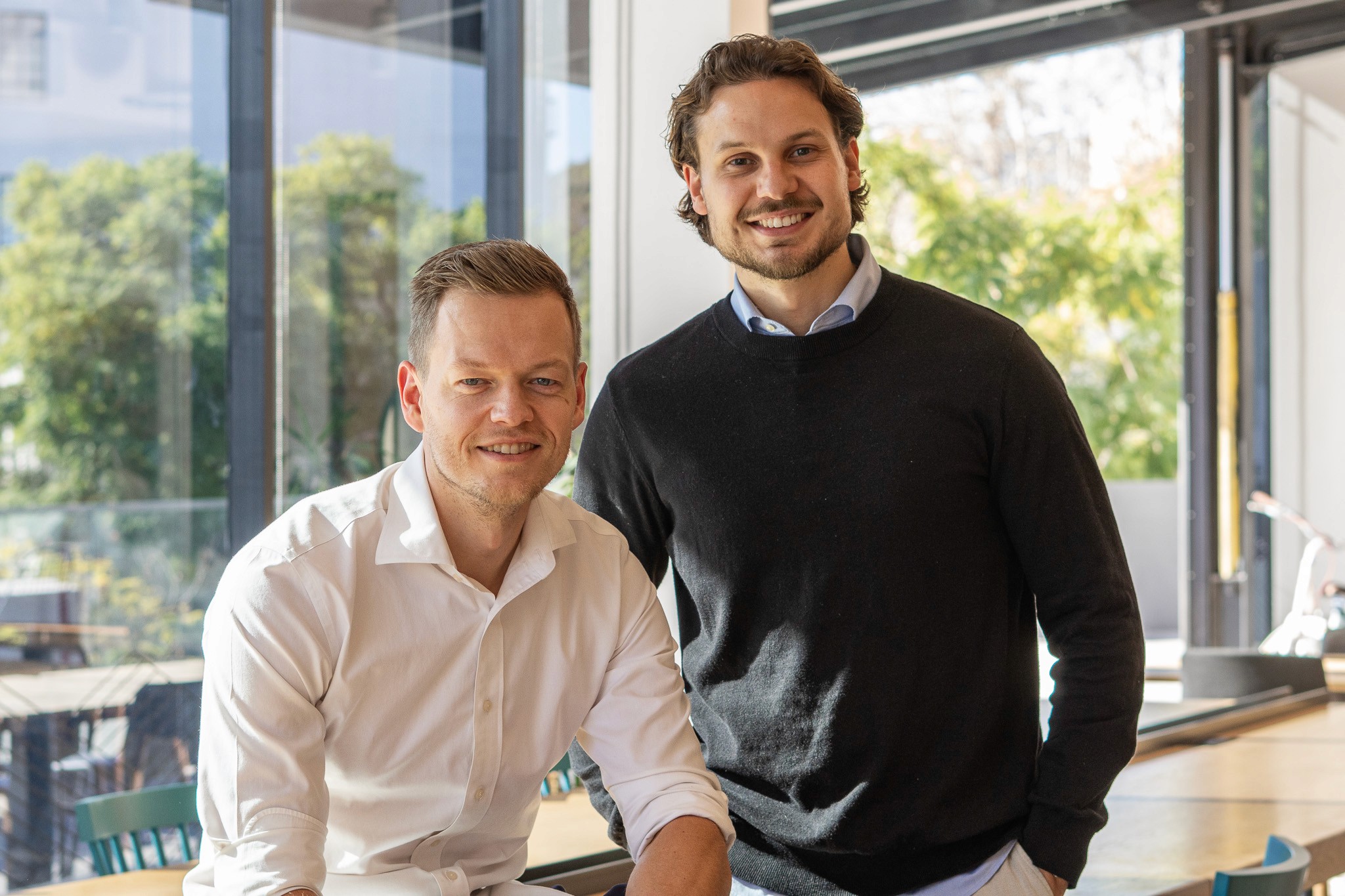 A young person with curly hair smiles at the camera while sitting in front of a laptop against a dark background.