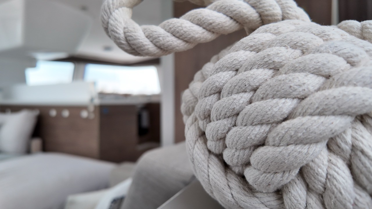 Macro close-up of a decorative white cotton monkey fist knot in the Bali 4.4 catamaran cockpit, with the bright modern saloon interior and walnut cabinetry softly blurred in the background, highlighting the nautical lifestyle detail of the yacht