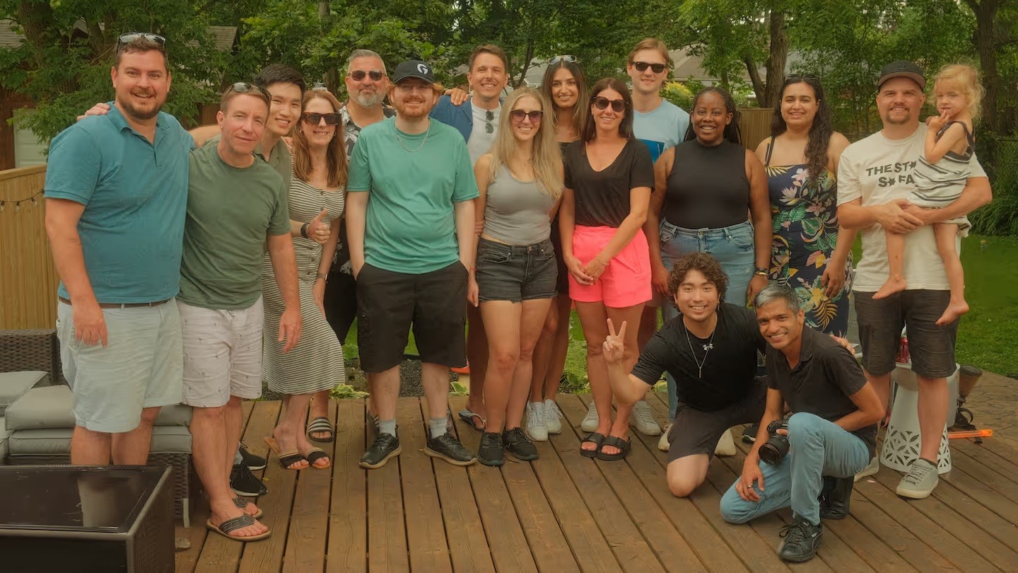 Group photo of around 18 people gathered on a wooden deck during a casual outdoor event, with a mix of men, women, and a child smiling at the camera. Most are dressed in summer attire, and the background shows a fenced yard with trees and greenery.