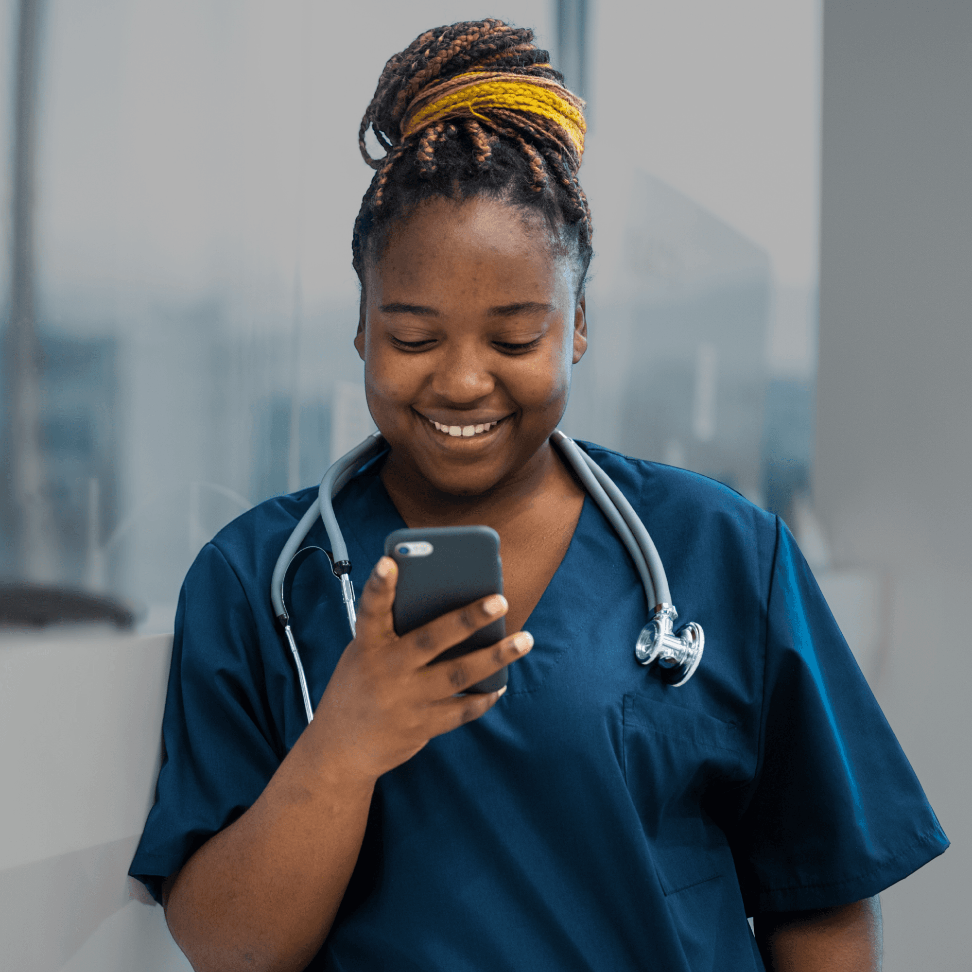 Black female health worker in blue scrubs looking at a phone and smiling, with a background showing a hospital setting.