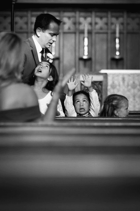 Documentary-style black and white image of a father with children in a church pew during a wedding, showcasing genuine family interaction and spontaneity in wedding photojournalism.