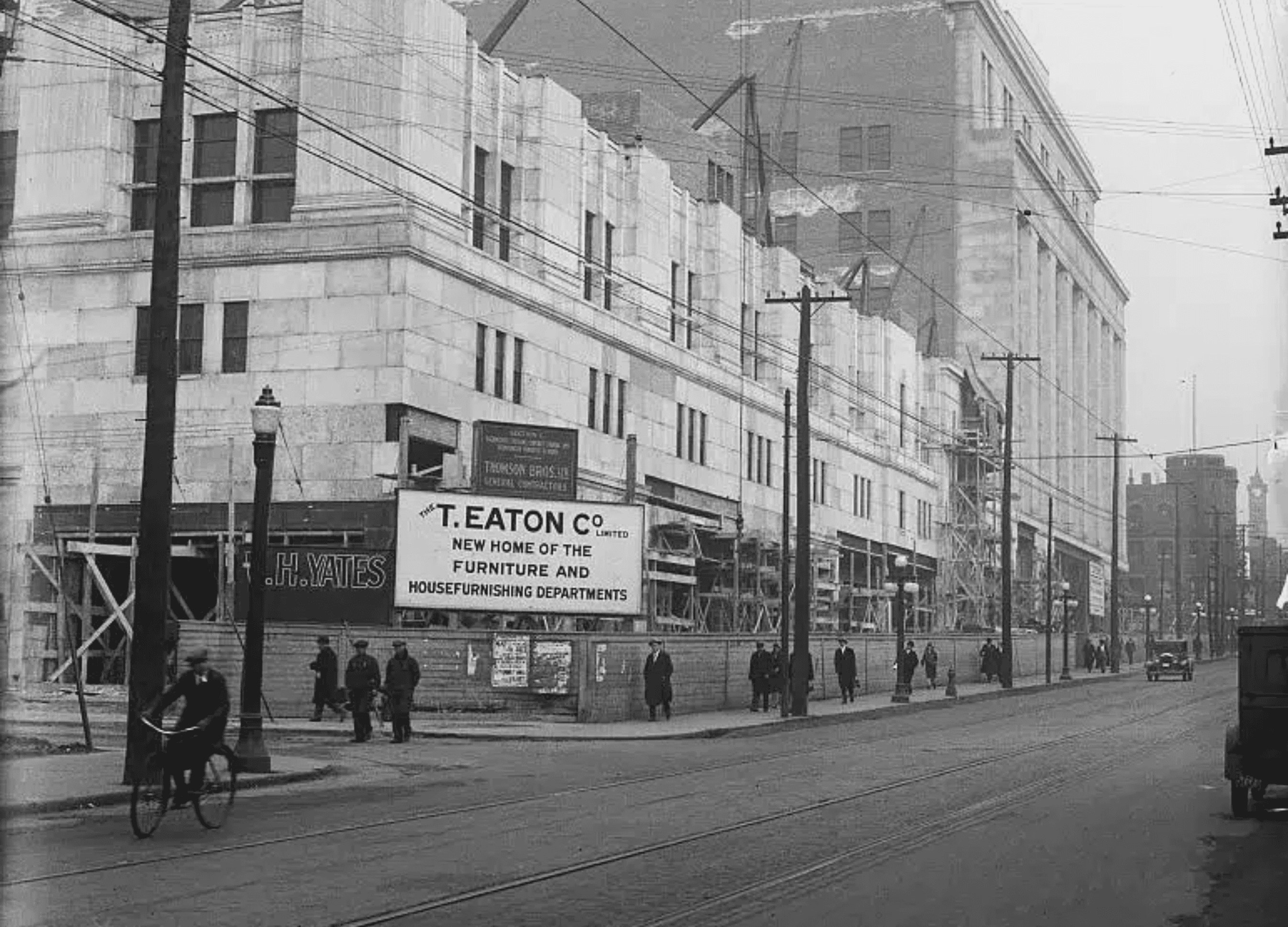 Historic black-and-white photograph of the T. Eaton Company building under construction, with workers and pedestrians on the street and a prominent sign announcing the new home of the furniture and housefurnishing departments. Power lines cross above and a cyclist rides along the sidewalk, reflecting a bustling city scene from the early 20th century.