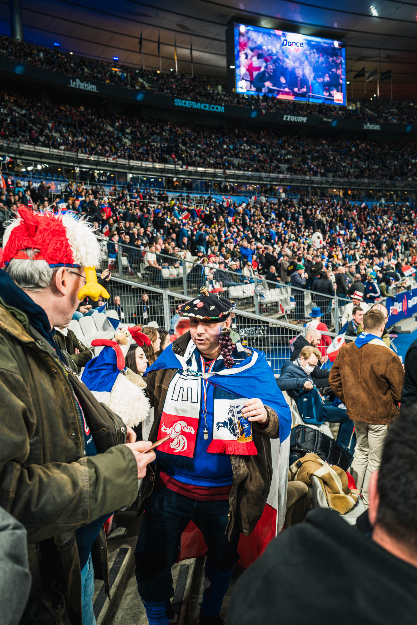 Un supporter français en béret couvert de pins, grappe de raisin à l'oreille et drapeau tricolore sur les épaules, une canette à la main, tribunes pleines derrière lui