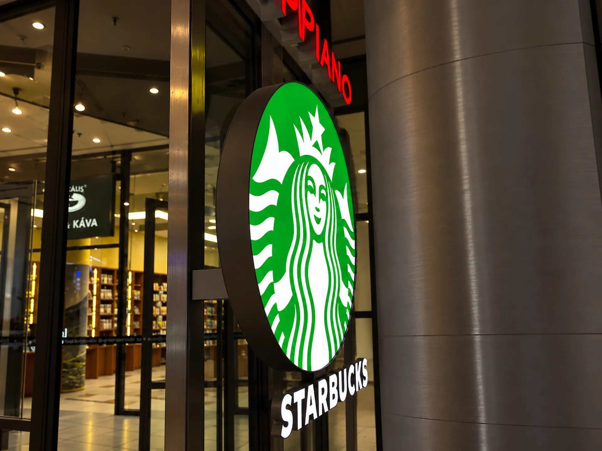 Kulaté světelné logo Starbucks se zelenou sirénou a bílý nápis na sloupu u vstupu do kavárny