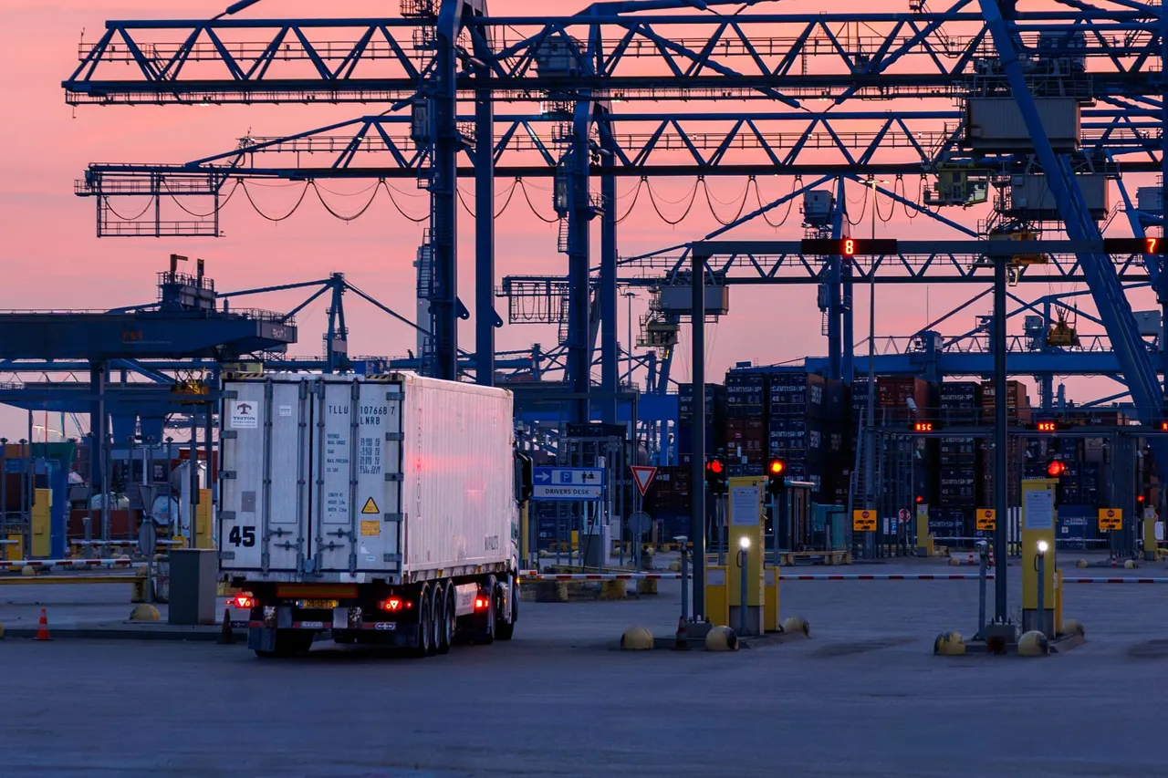 A refrigerated container truck crossing a terminal under cranes at dusk