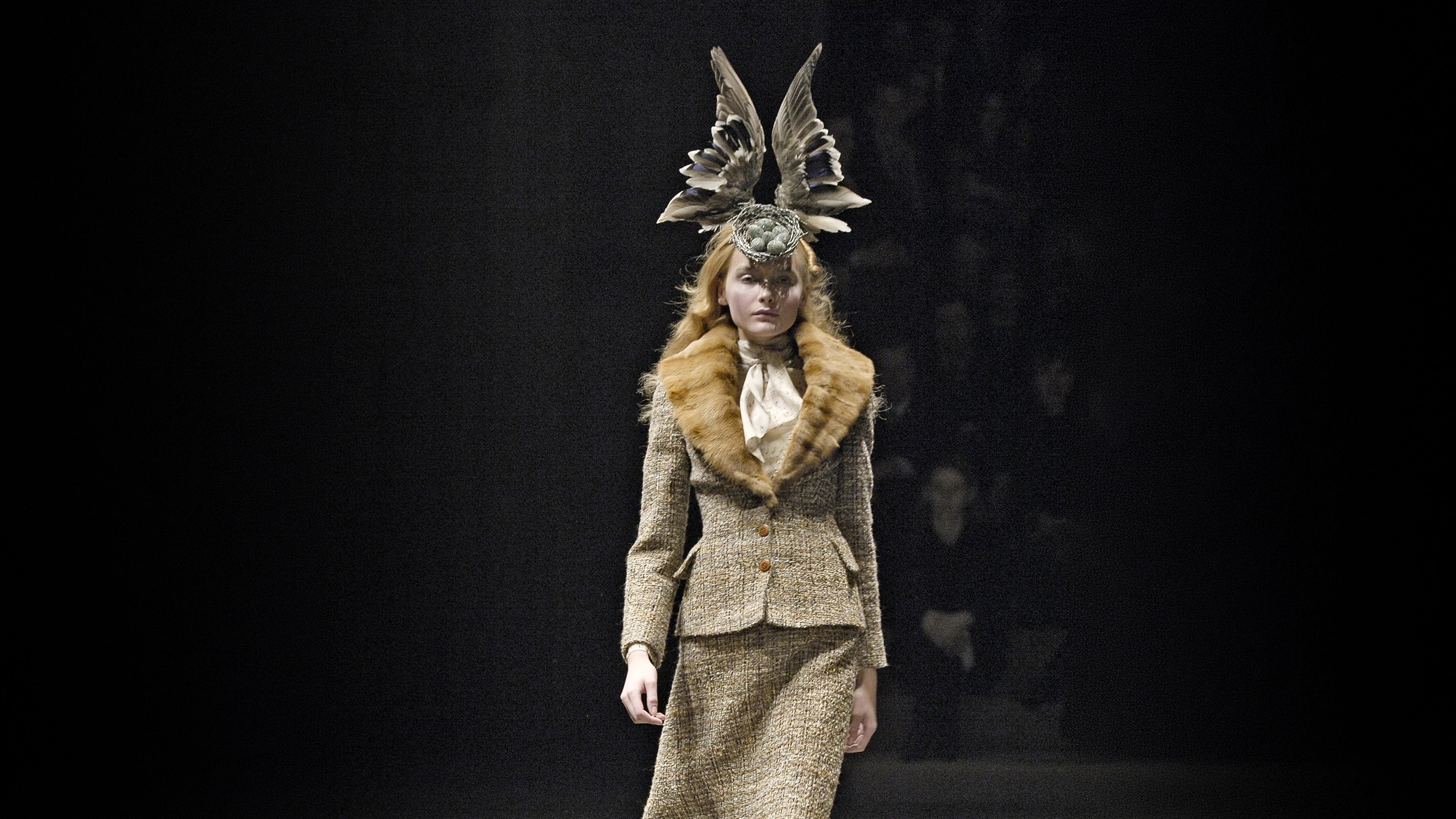 Model on a dark runway wearing a Shaun Leane avant-garde bird's nest headpiece featuring two realistic upright bird wings and small blue eggs, paired with a tweed jacket and fur collar.