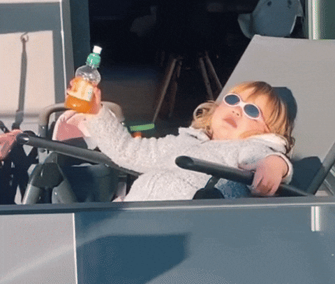 A toddler sprawled in a stroller wearing sunglasses, sipping juice with one arm outstretched like a cool driver.