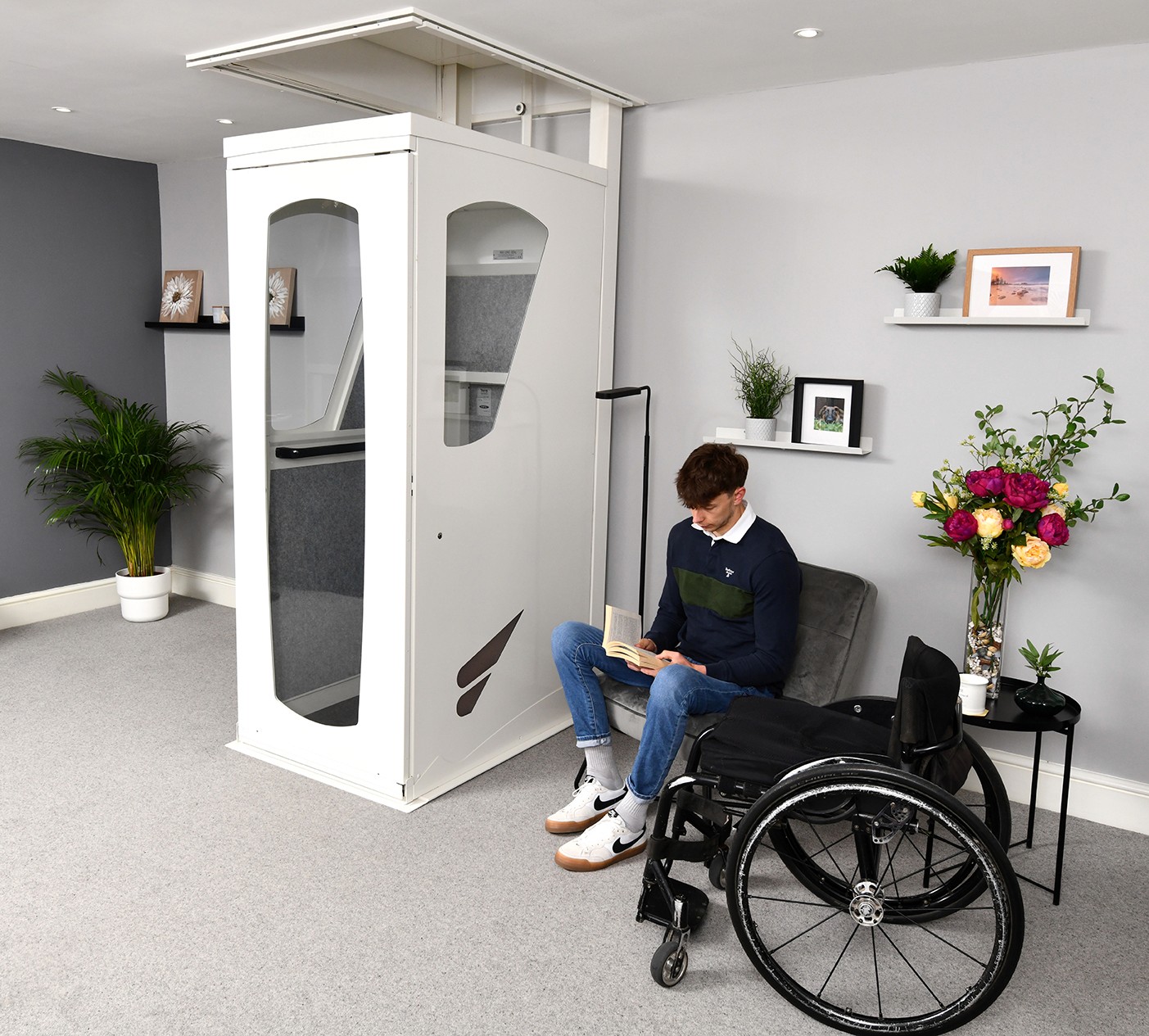 Wheelchair user seated beside an Elevate AVA Reach through-floor homelift installed in a residential living room, with white cabin doors open at ground floor level