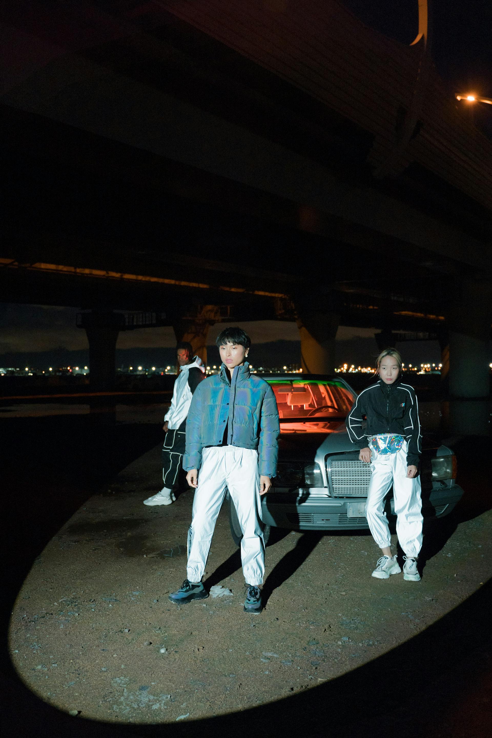 Three people in reflective streetwear pose at night in front of a vintage car parked under a large bridge.
