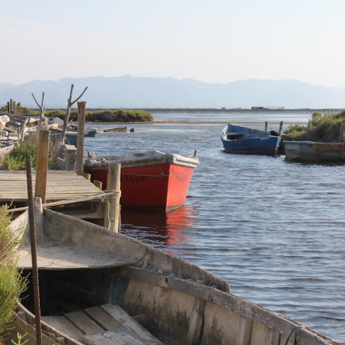 Pequeñas embarcaciones de madera atracadas a lo largo de un muelle de madera en un cuerpo de agua tranquilo, con montañas visibles al fondo.