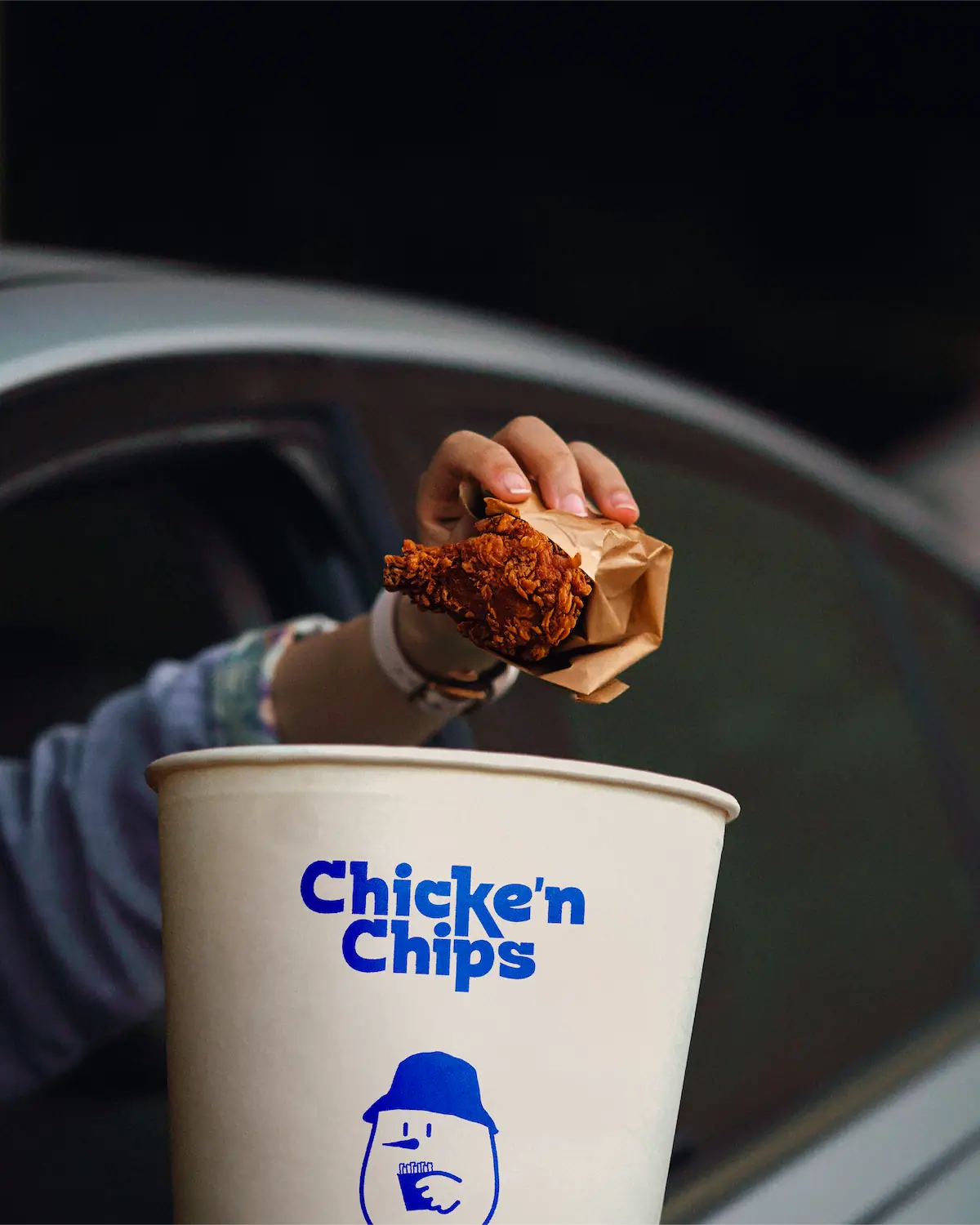 Main tenant un tender de poulet frit au-dessus d'un gobelet Chicke'n Chips brandé bleu et blanc, depuis une fenêtre de voiture.