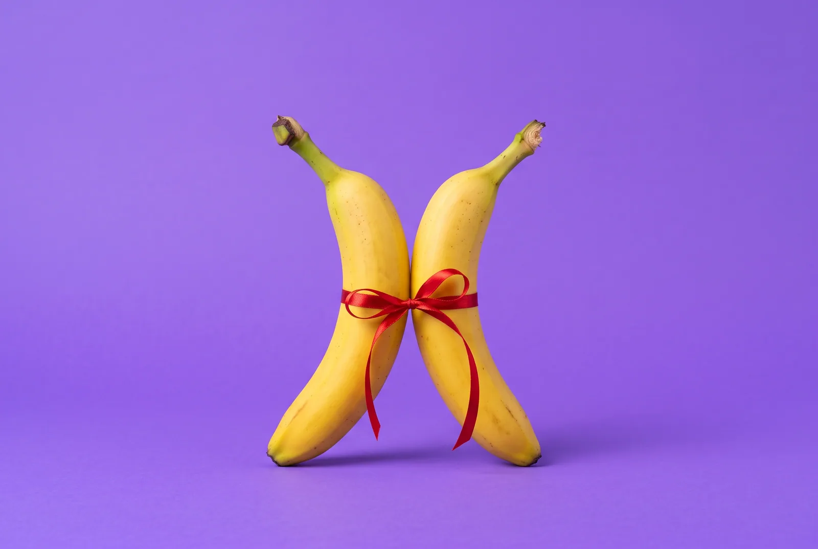 Accountability partner for porn concept: two bananas standing side by side, loosely tied together with a red ribbon on a lilac backdrop