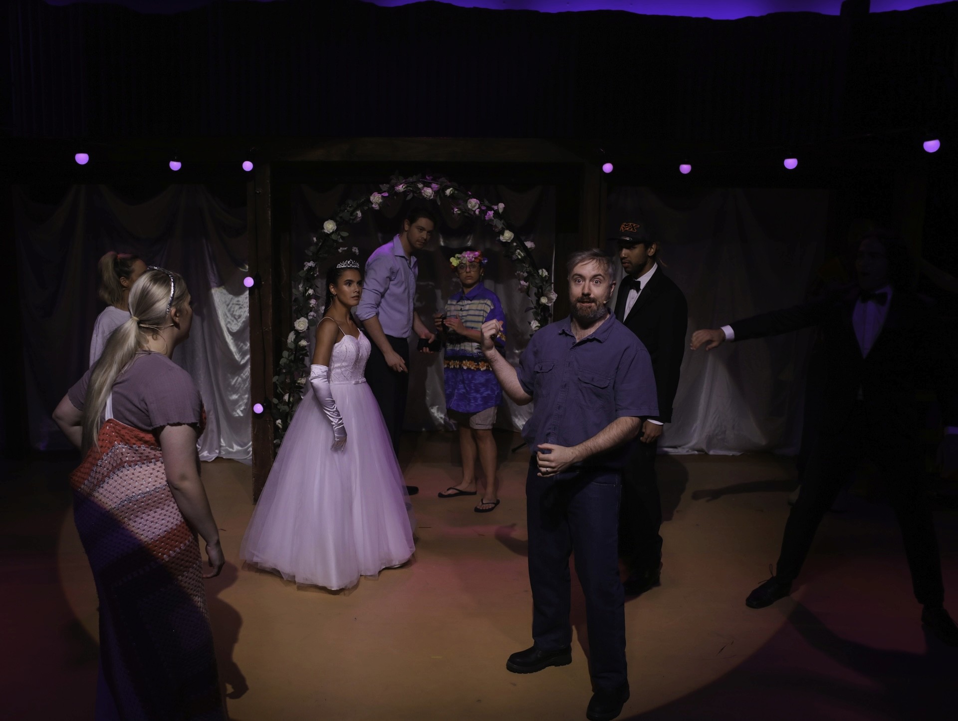 A wedding scene unfolds on stage with performers arranged around a decorated floral arch. A performer in a white ballgown stands at the centre, while others in colourful costumes react to the moment. A man in casual clothes stands foreground, mid-speech, breaking the fourth wall. The lighting is soft and theatrical.