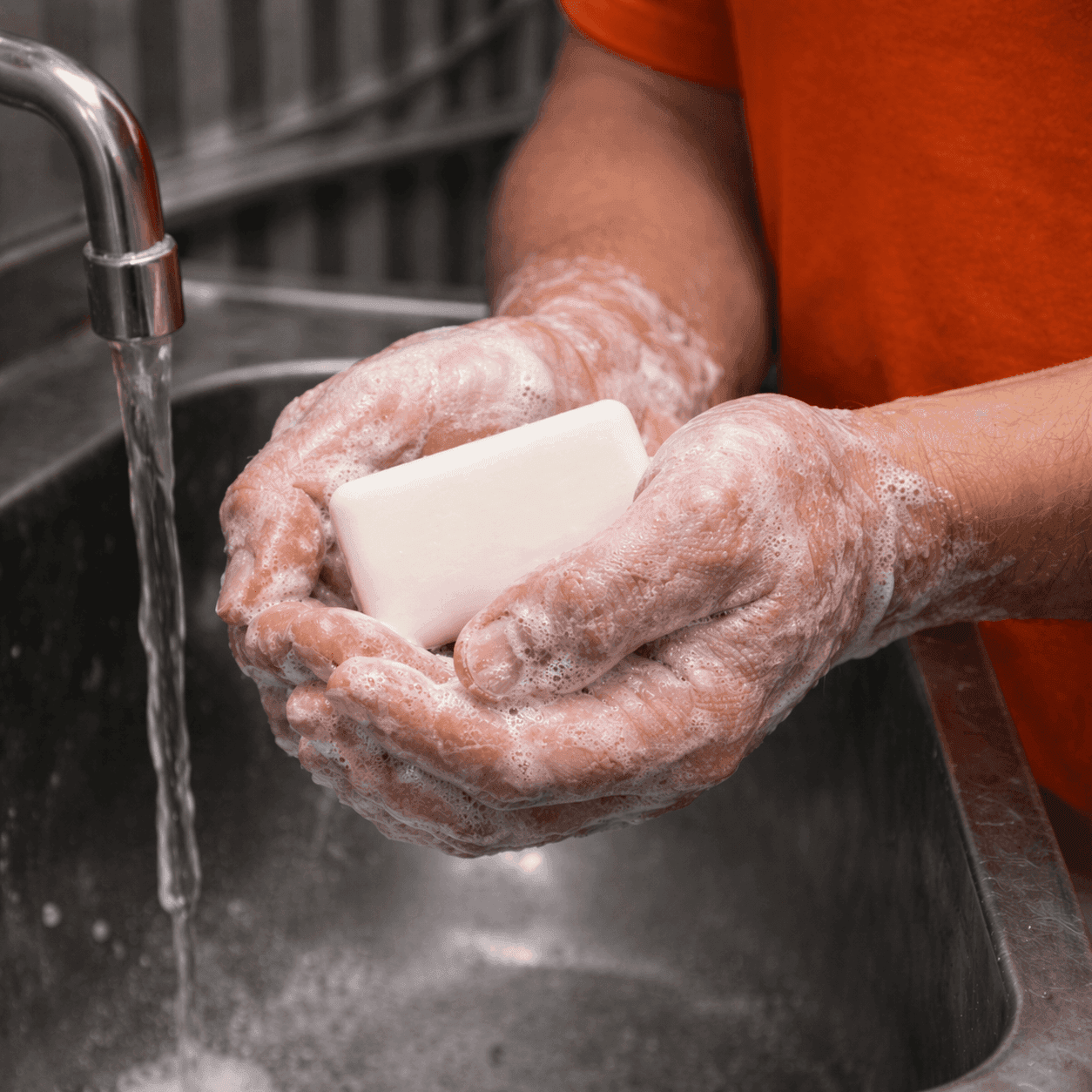 Person washing hands with Good Day soap bar by Transmacro Amenities in a prison sink with running water and visible foam, close-up lifestyle usage scene