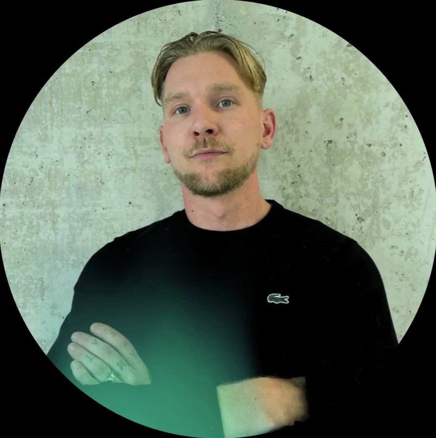 A man with light hair stands confidently, arms crossed, against a circular, softly glowing background.