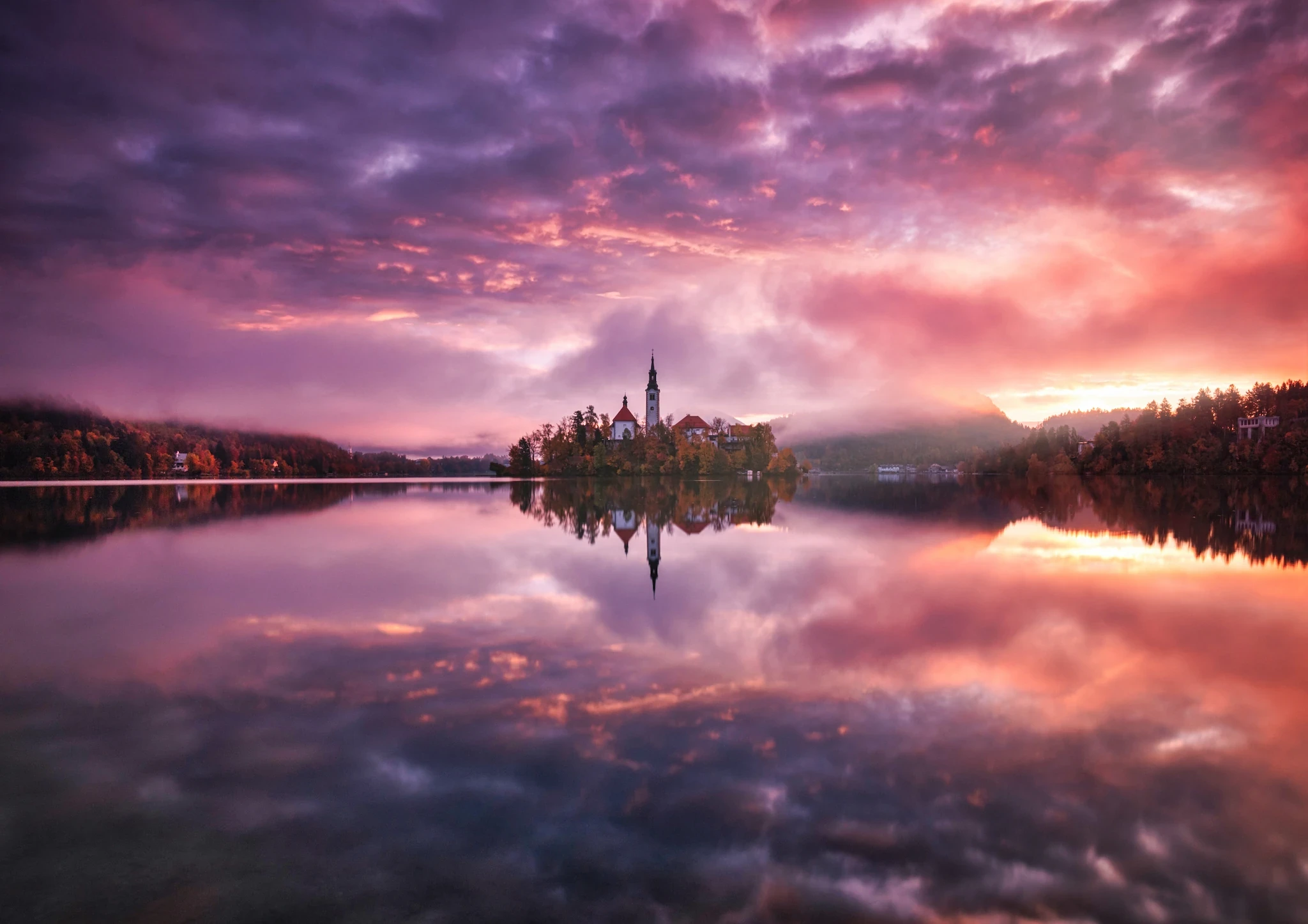 The island church at Lake Bled, Slovenia, reflected in calm water under a vibrant purple and orange sunrise.