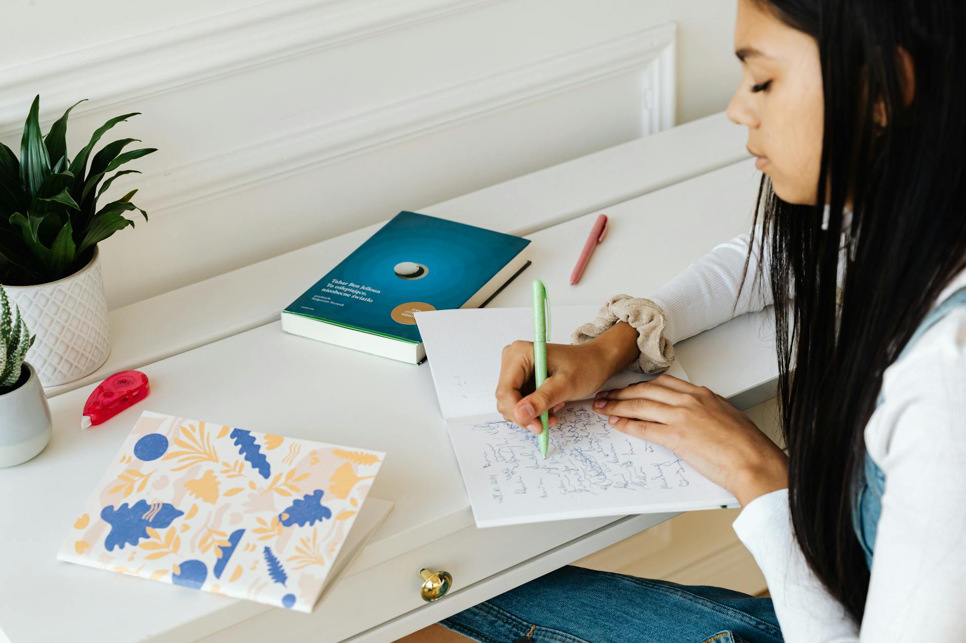 A young student pointing at words in a phonics workbook while reading a book at a wooden school desk.