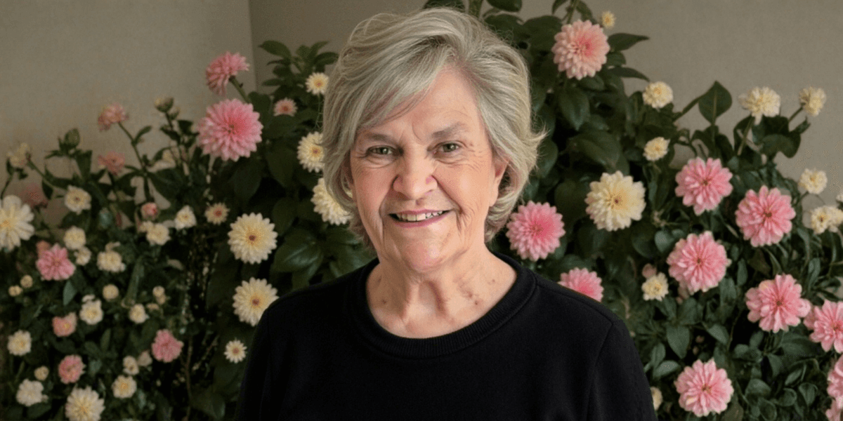 Smiling older woman with gray hair stands before pink and white flowers, representing psychic Barbara Rae and her intuitive guidance.