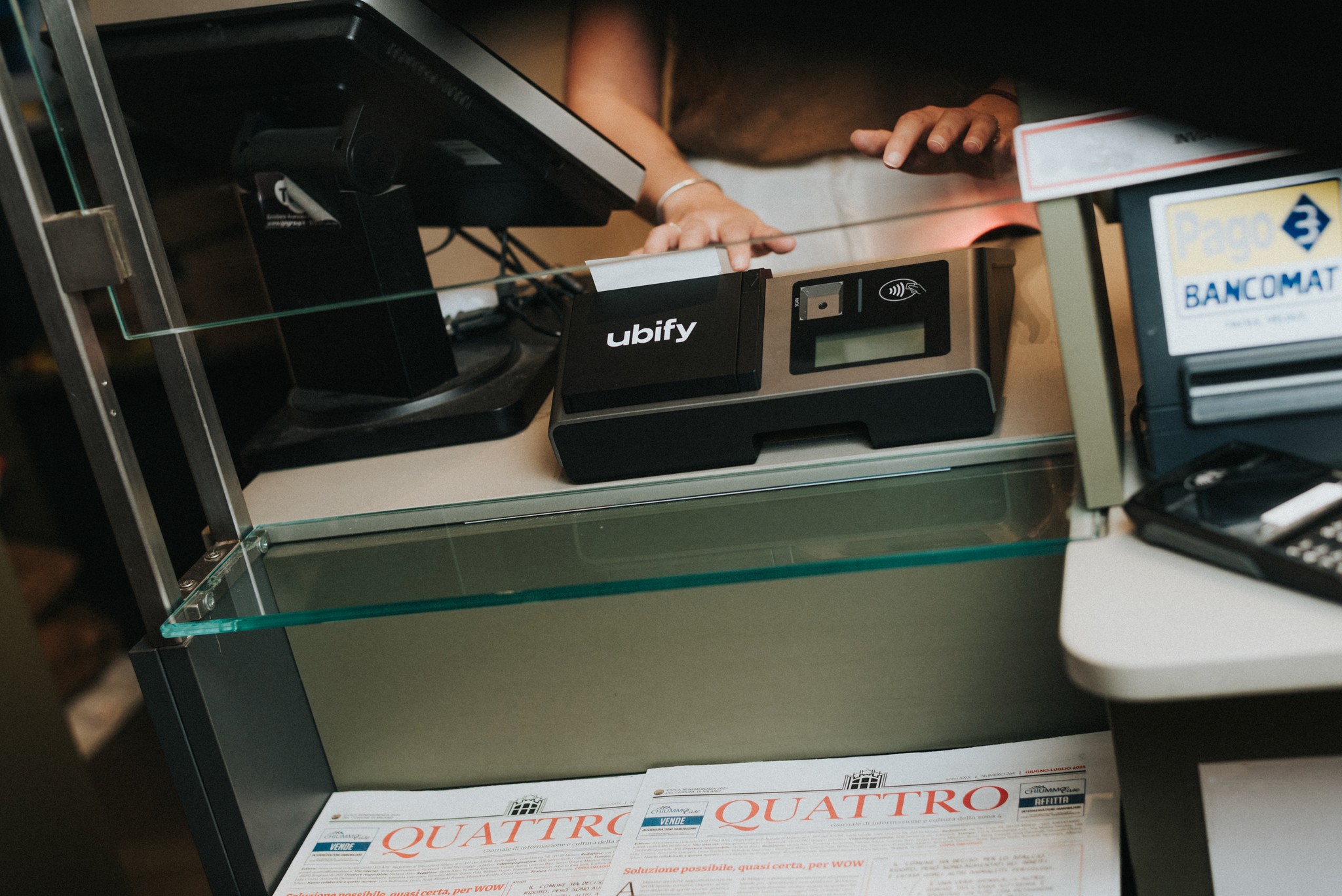 Cassa Ubify su bancone di tabaccheria, affiancata da terminale POS e giornali esposti sotto al vetro.
