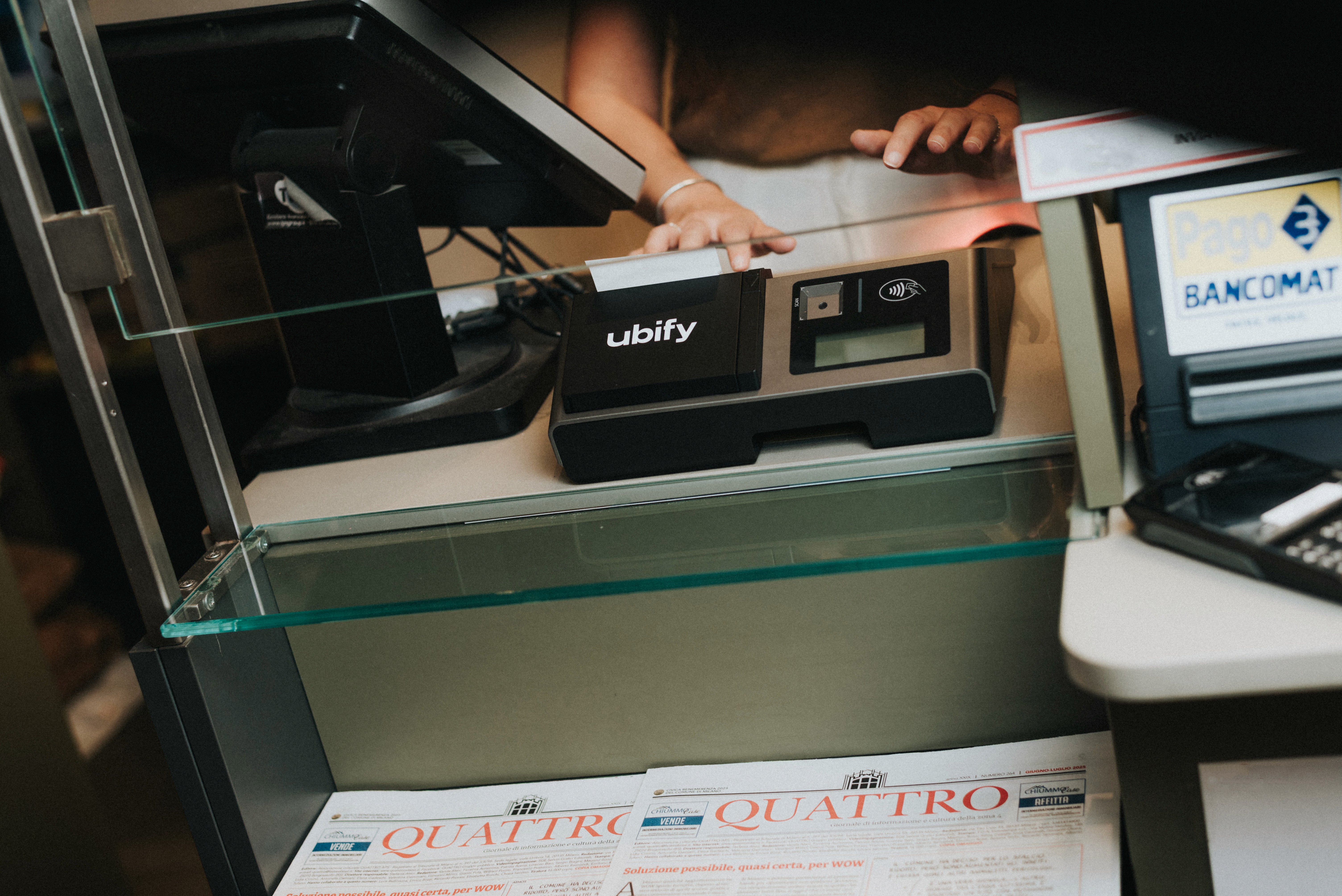 Cassa Ubify su bancone di tabaccheria, affiancata da terminale POS e giornali esposti sotto al vetro.