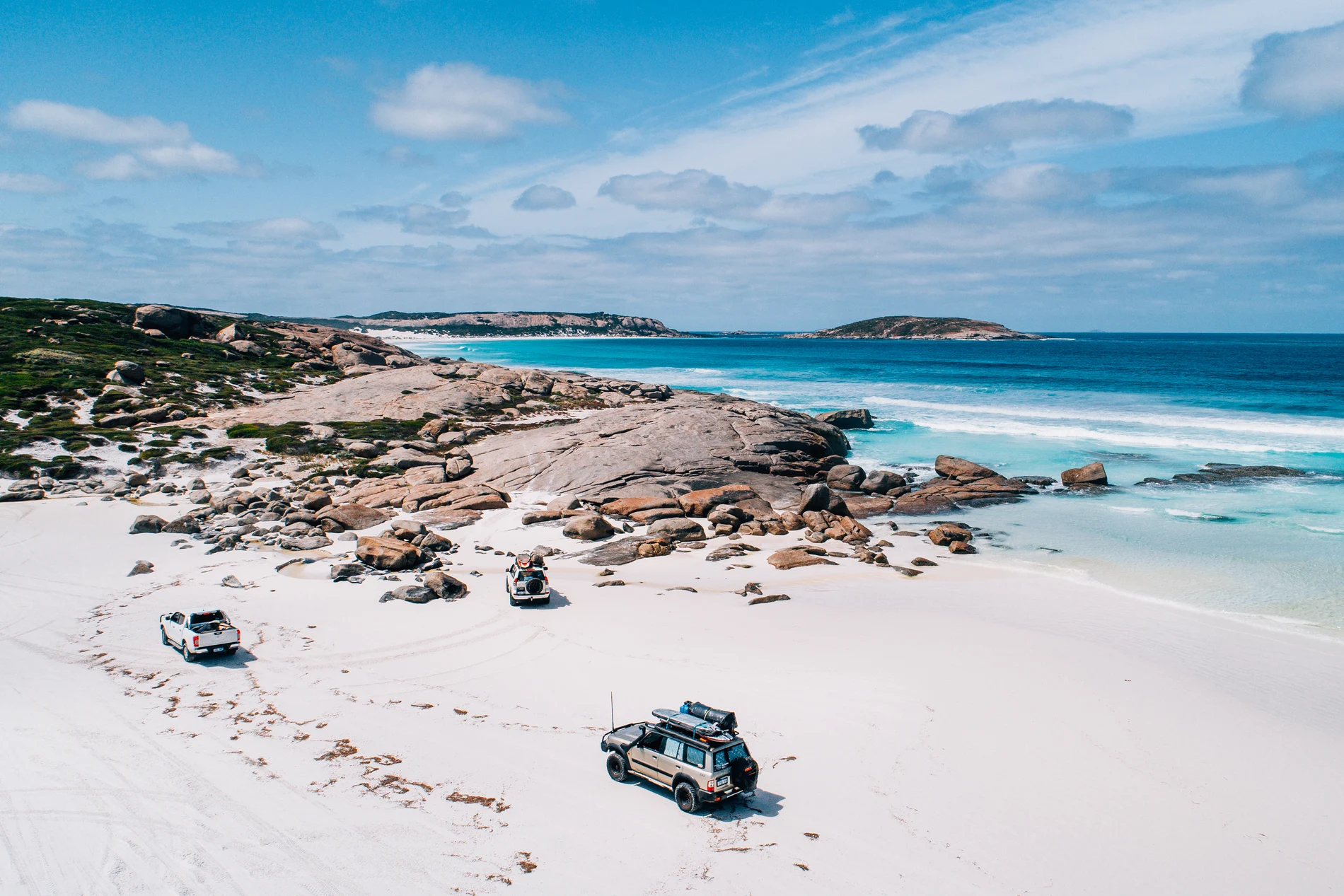 Four-wheel drives parked on a white beach beside granite boulders and turquoise sea