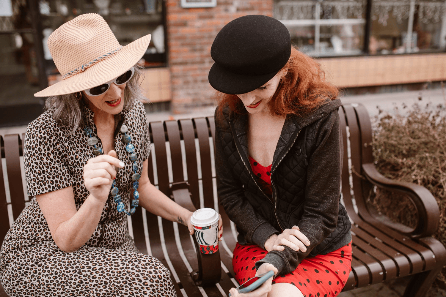 Two older women sitting on a bench outside a shop with their coffees, laughing over something one of them is holding.