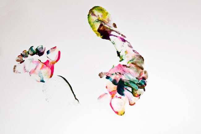 The image features abstract watercolor paintings on a white background. On the left side, there is a cluster of pink, orange, and white shapes that resemble a flower or head. To the right, a vertical splash of green, yellow, and white paint extends upward, resembling a splash or burst. The bottom left corner contains a small, dark splash or stroke. The colors are vibrant and fluid, typical of watercolor art.