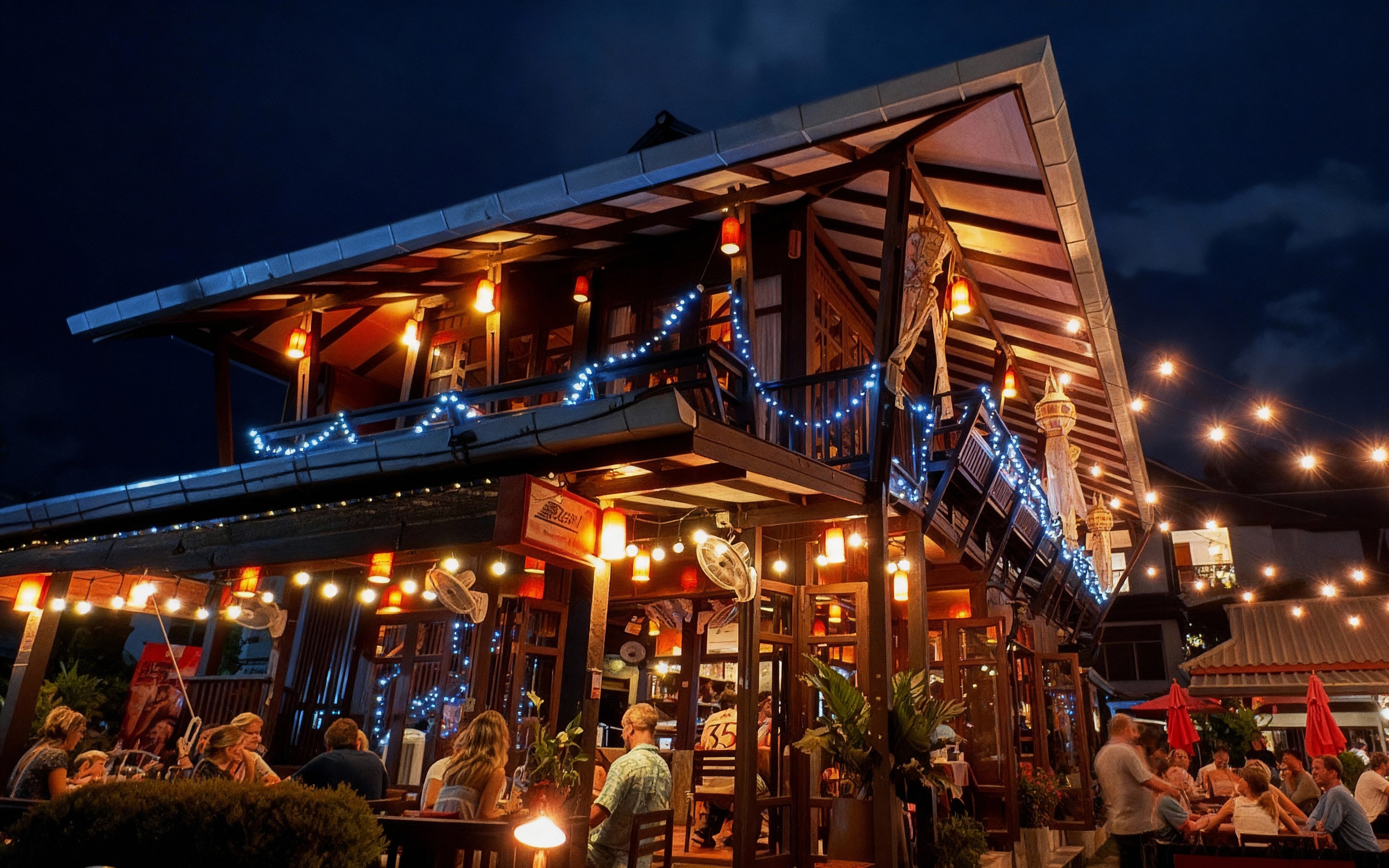 Warmly lit two-story teakwood restaurant filled with diners under string lights and lanterns, showcasing Dash Restaurant & Bar’s cozy evening atmosphere in Chiang Mai’s Old City.