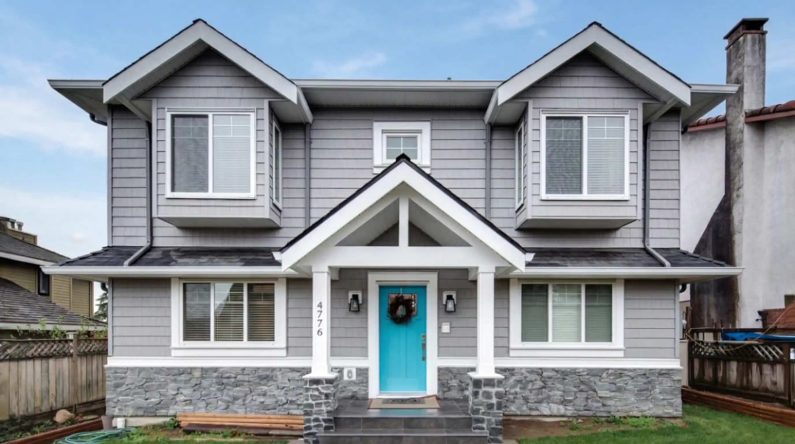 Modern two-story custom home exterior in Burnaby featuring gray horizontal siding, natural stone foundation accents, contemporary architectural design with turquoise blue front door, and multi-generational layout built by Vancouver Custom Homes
