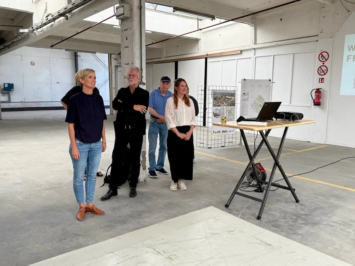 Personengruppe bei einer Präsentation in einer großen Industriehalle; Laptop und Beamer auf einem Stehtisch, offene Raumstruktur mit Betonstützen.