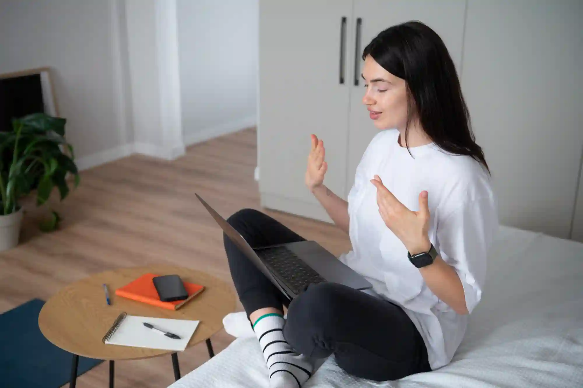 A woman sits cross-legged on a bed, actively gesturing during a professional video call on her laptop.