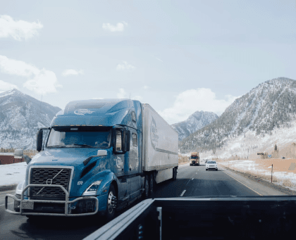 Semi-truck driving on a highway through mountainous terrain during winter conditions.