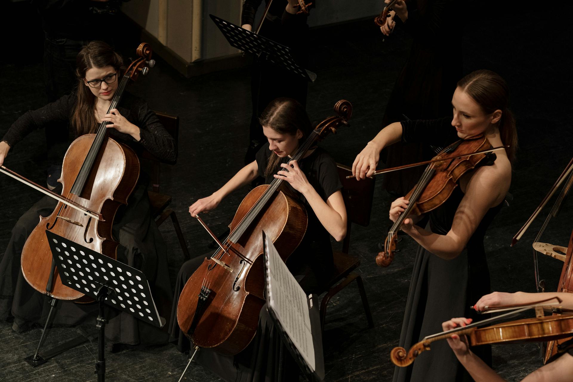 A high-angle shot of an orchestra section featuring three women in black formal attire playing two cellos and a violin on a dark stage.