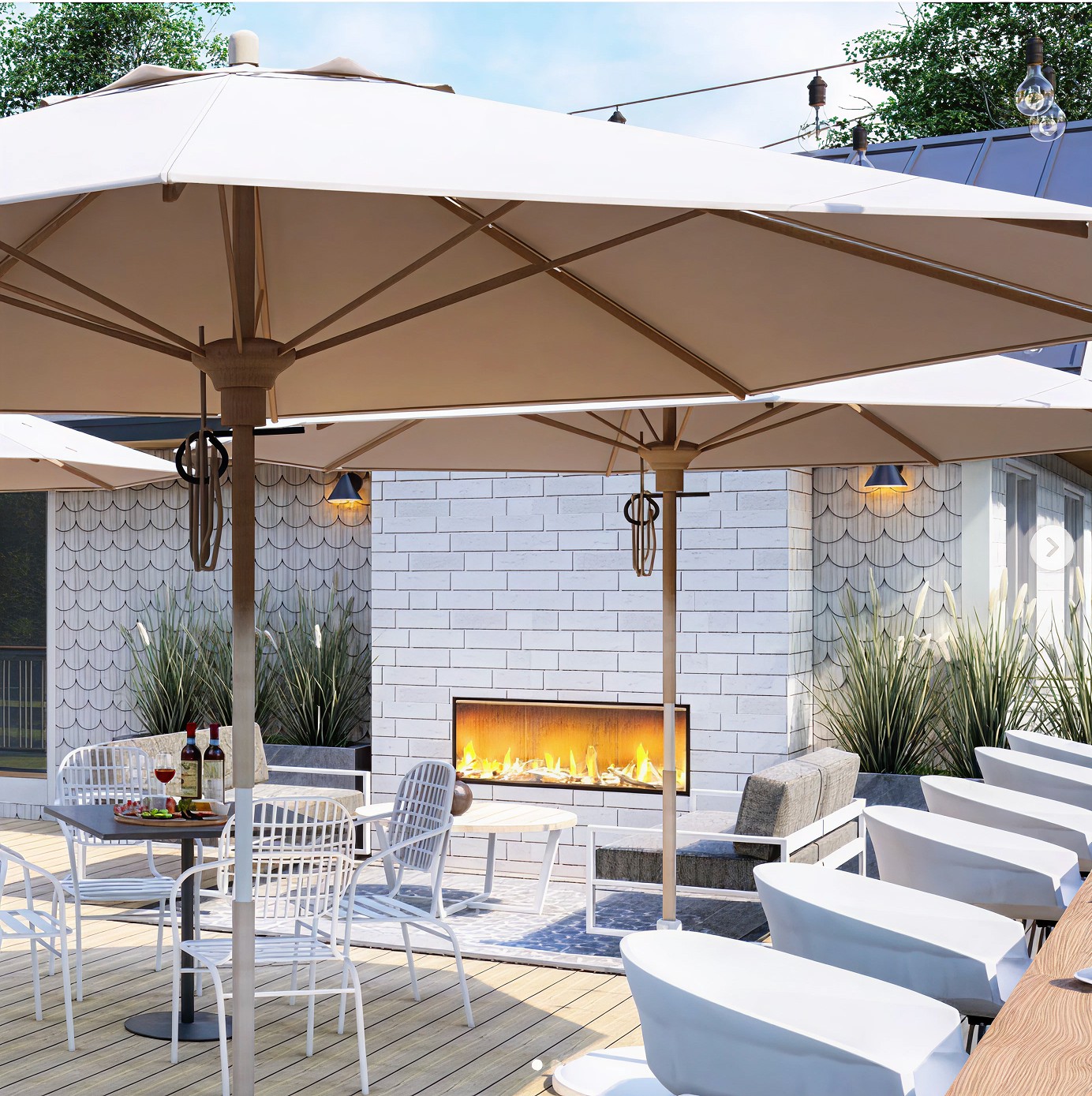 Outdoor patio space at a boutique motel featuring large cream-colored umbrellas, a long modern fireplace built into a white brick wall, white patio furniture, and a cozy atmosphere perfect for lounging or dining al fresco.