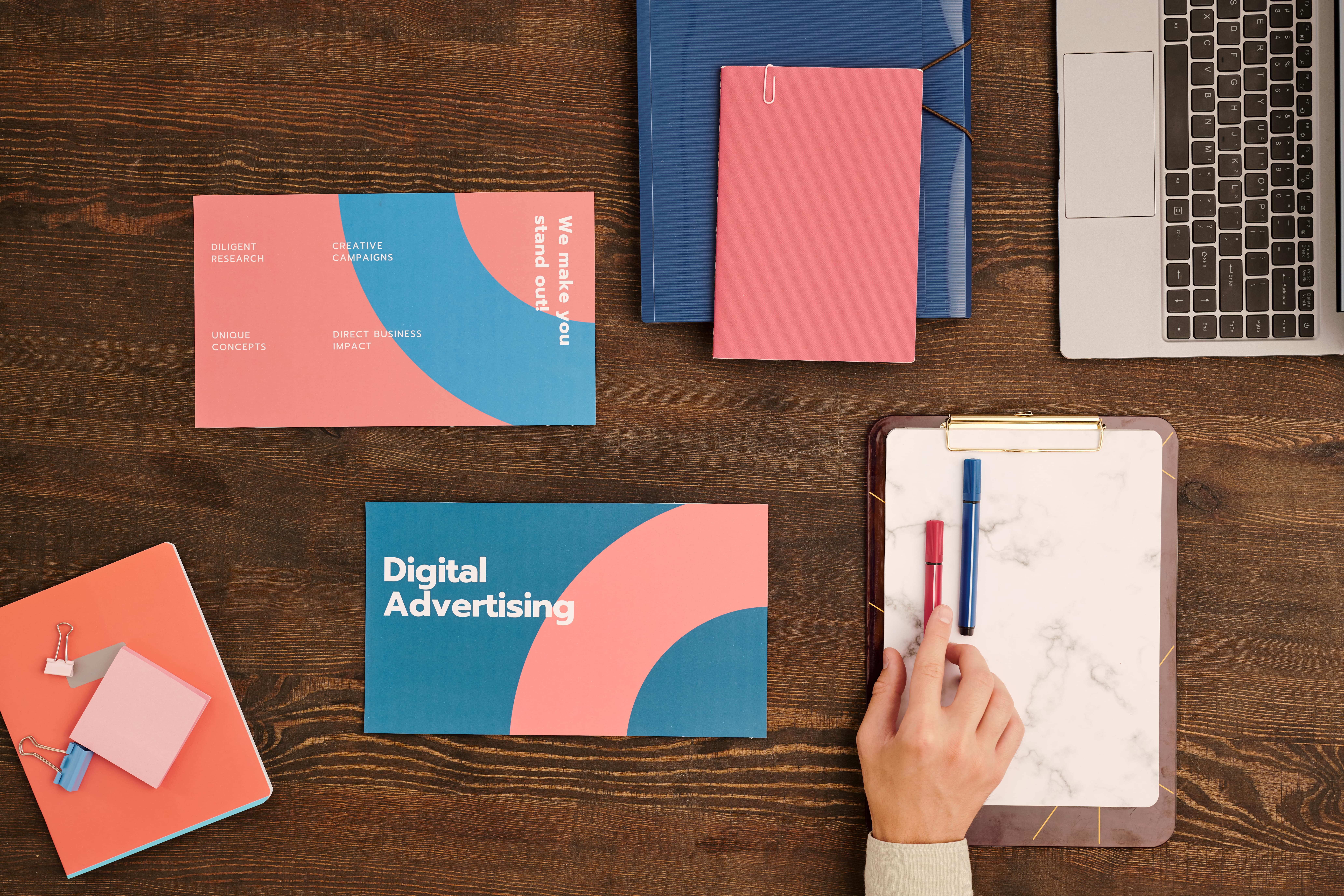 Flat lay of digital advertising materials, notebooks, and a laptop on a wooden desk, with a hand reaching for a pen—representing modern marketing strategy and creative planning.